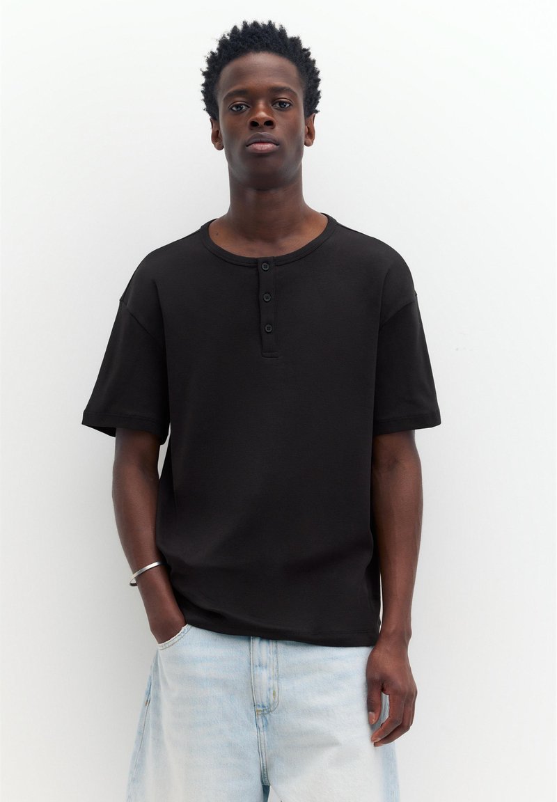 Jeune homme aux cheveux courts et bouclés, portant un t-shirt henley noir à manches courtes et un jean bleu clair, posant devant un fond blanc uni.