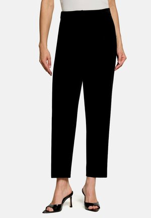 Femme portant un pantalon noir taille haute droit associé à des chaussures à talons hauts noires ouvertes à l’orteil, visible de la taille aux pieds.