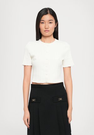 Mujer con cabello negro lacio, vistiendo una camiseta corta de manga corta blanca con botones y una falda negra de cintura alta con detalles decorativos en dorado.