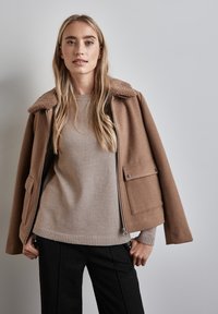 Braune Wollmischungjacke mit strukturiertem Kragen, zwei Fronttaschen und einem Metallreißverschluss, getragen über einem beigen Strickpullover und schwarzen Hosen.