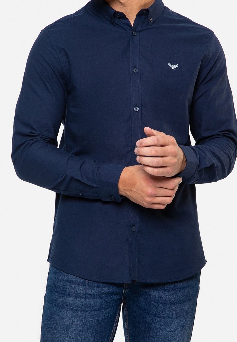 Camisa de botones de manga larga en azul oscuro, hecha de una tela suave, con un diseño ajustado y un pequeño emblema bordado en el pecho.