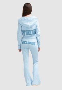 Survêtement bleu clair comprenant une veste à capuche courte et un pantalon évasé, avec "TRUE" et "RELIGION" imprimés en lettres audacieuses et contrastées.