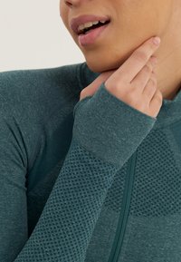 Langärmliges Sportoberteil in dunkeltürkis mit strukturierten Mesh-Akzenten, einem Frontreißverschluss und enganliegenden Ärmeln mit Daumenlöchern für zusätzlichen Schutz.