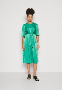 Grünes Satin-Kleid mit kurzen Puffärmeln, einem gebundenen Taillendetail und einem mittellangen Rock. Kombiniert mit schwarzen offenen Mules.