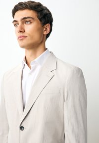 Junger Mann mit kurzem dunklem Haar, der einen hellbeigen, strukturierten Blazer und ein weißes Hemd mit Kragen trägt, schaut leicht nach links vor weißem Hintergrund.