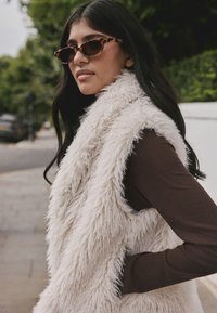 Kunstpelzweste in Creme mit strukturiertem Finish, getragen über einem braunen Langarmshirt. Cateye-Sonnenbrille mit Schildpatt-Muster.