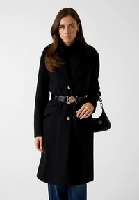 Črni plašč iz volnene mešanice z ovratnikom iz umetnega krzna, zapenjanjem na gumbe, dvema sprednjima žepoma in usklajenim pasom, kombiniran z črno torbico.