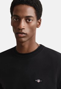 Zwarte trui met ronde hals, voorzien van een klein logo op de linkerborst. Gladde textuur en een aansluitend ontwerp.