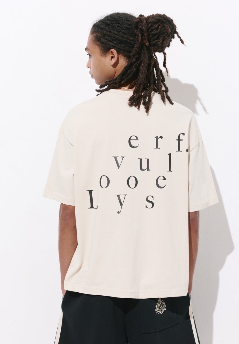 Personne avec des dreadlocks portant un T-shirt beige oversize avec des lettres noires dispersées et un pantalon noir avec un petit emblème.