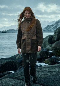 Bruine gewatteerde jas met fleece mouwen, contrasterende texturen en drukknoopsluiting, in combinatie met donkere broek en laarzen, tegen een rotsachtige achtergrond.