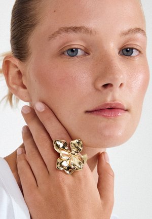 Close-up van een persoon met een lichte huid en blauwe ogen die een grote, glanzende gouden abstracte bloemvormige ring draagt op de hand vlak bij het gezicht.