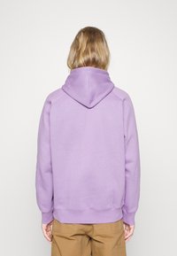 Persona con capelli biondi di lunghezza spalle indossa una felpa semplice color lavanda e pantaloni marroni, voltata di spalle contro uno sfondo bianco.