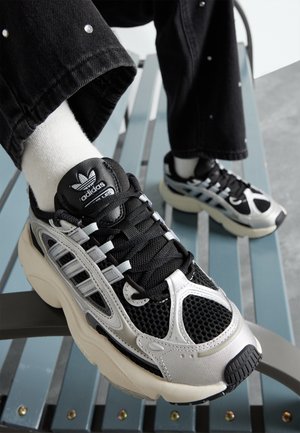 Detalje af fodtøj, der viser sorte og sølvfarvede Adidas-sneakers med hvide sokker og sorte bukser, hvilende på en lyseblå afskåret bænk.