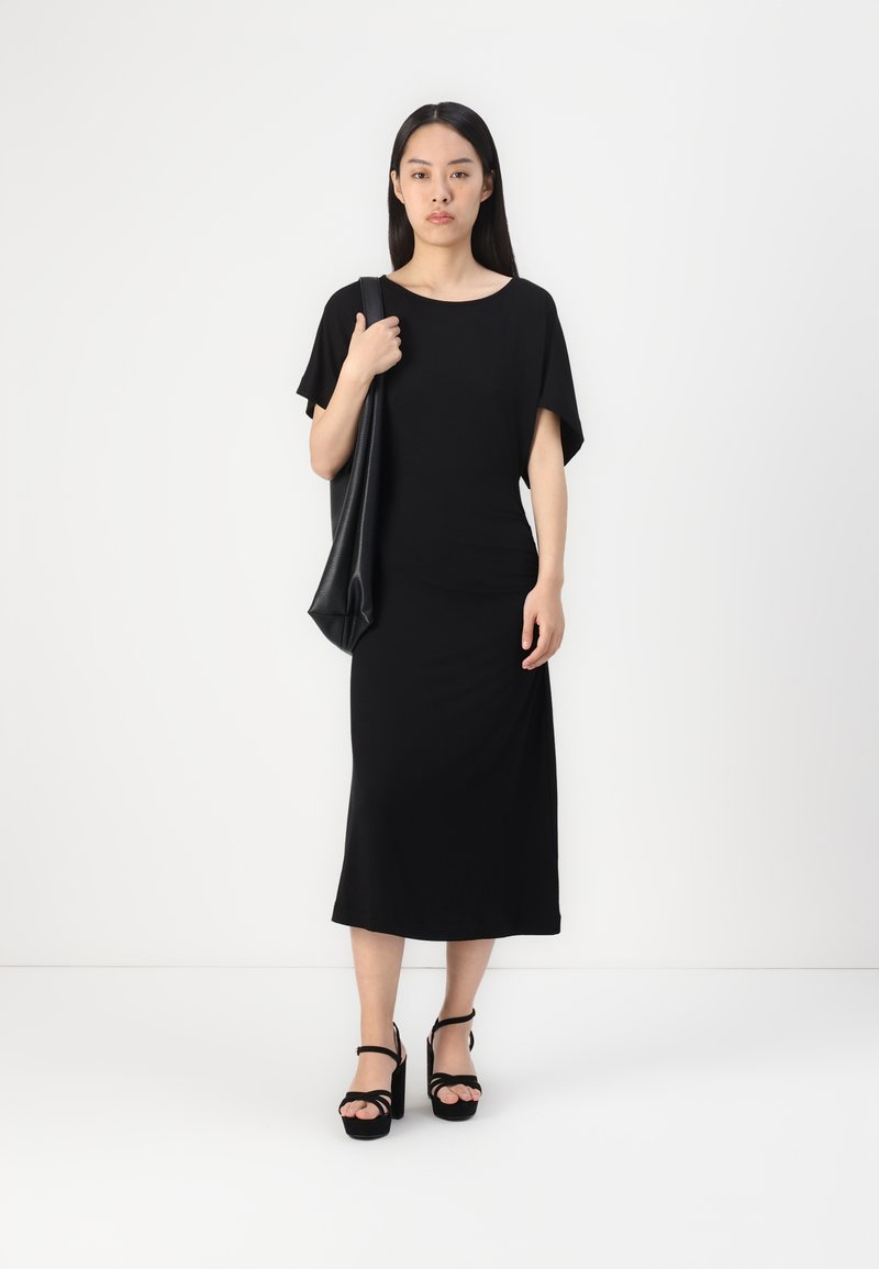 Zwarte, losse jurk met korte mouwen, knielange ontwerp en een gladde textuur. Gepaarde met zwarte sandaaltjes met hoge hakken en een schoudertas.