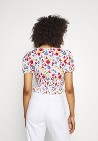 Top corto floreale con schiena arricciata, maniche a sbuffo corte e colori vivaci su uno sfondo chiaro, abbinato a pantaloni bianchi a vita alta.