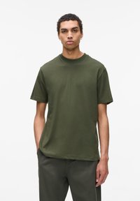 Tricou din bumbac verde măsliniu, cu mânecă scurtă, guler rotund, croială lejeră, textură netedă, design minimalist, asortat cu pantaloni verzi corespunzători.