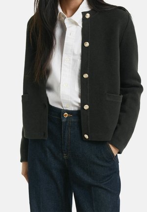 Frau trägt eine schwarze, geknöpfte Strickjacke über einem weißen Hemd mit Kragen, kombiniert mit dunkelblauen, hoch taillierten Jeans, eine Hand in der Tasche.
