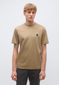 Bézs, rövid ujjú pamut póló, kerek nyakkivágással és egy kis fekete logóval a bal mellrészen. Egyszerű, egyenes szabás.