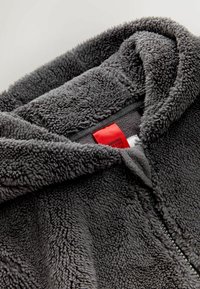 Grå fleece-hoodie med en blød, tekstureret overflade. Har et rødt mærke nær lynlåsen og en hætte med en rund form.