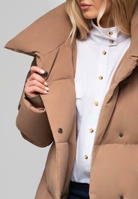 Brun pufferjakke med oversized krave, der har sorte knapper og en glat tekstur, lagt over en hvid skjorte med runde gyldne knapper.