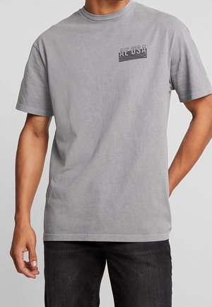 Homme portant un t-shirt gris clair à manches courtes avec le texte « AE USA » et un logo rayé sur la poitrine gauche, associé à un jean noir.