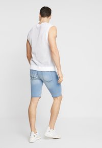 Top blanco sin mangas de malla con un corte relajado, combinado con pantalones cortos de mezclilla azul claro. La modelo lleva zapatillas blancas. Conjunto sencillo y casual.