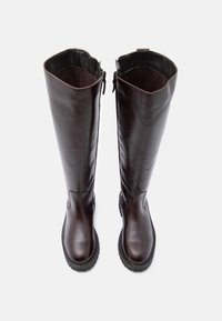 Um par de botas altas, de couro castanho escuro, com fechos laterais e solas grossas de borracha preta, exibidas de cima sobre um fundo branco.