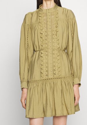 Femme portant une robe à manches longues vert olive clair avec un devant en dentelle boutonnée, une taille cintrée et un ourlet de jupe froncé.