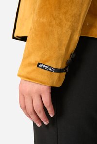 Geltonos veliūrinės striukės rankovė su etikete, ant rankogalių yra juodi mygtukai. Audinys yra minkštas, o dizainas prigludęs.