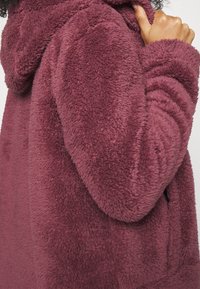Polarowa bluza w głębokim burgundzie, z teksturowanym, plush materiałem, kapturem oraz ribbowymi detalami na mankietach, zapewniającymi dodatkowy komfort.