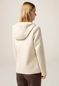 Beige Wolle-Mischung Kapuzenjacke mit glatter Textur, tailliertem Design und Nahtdetail auf der Rückseite, kombiniert mit dunkelbraunen Hosen.