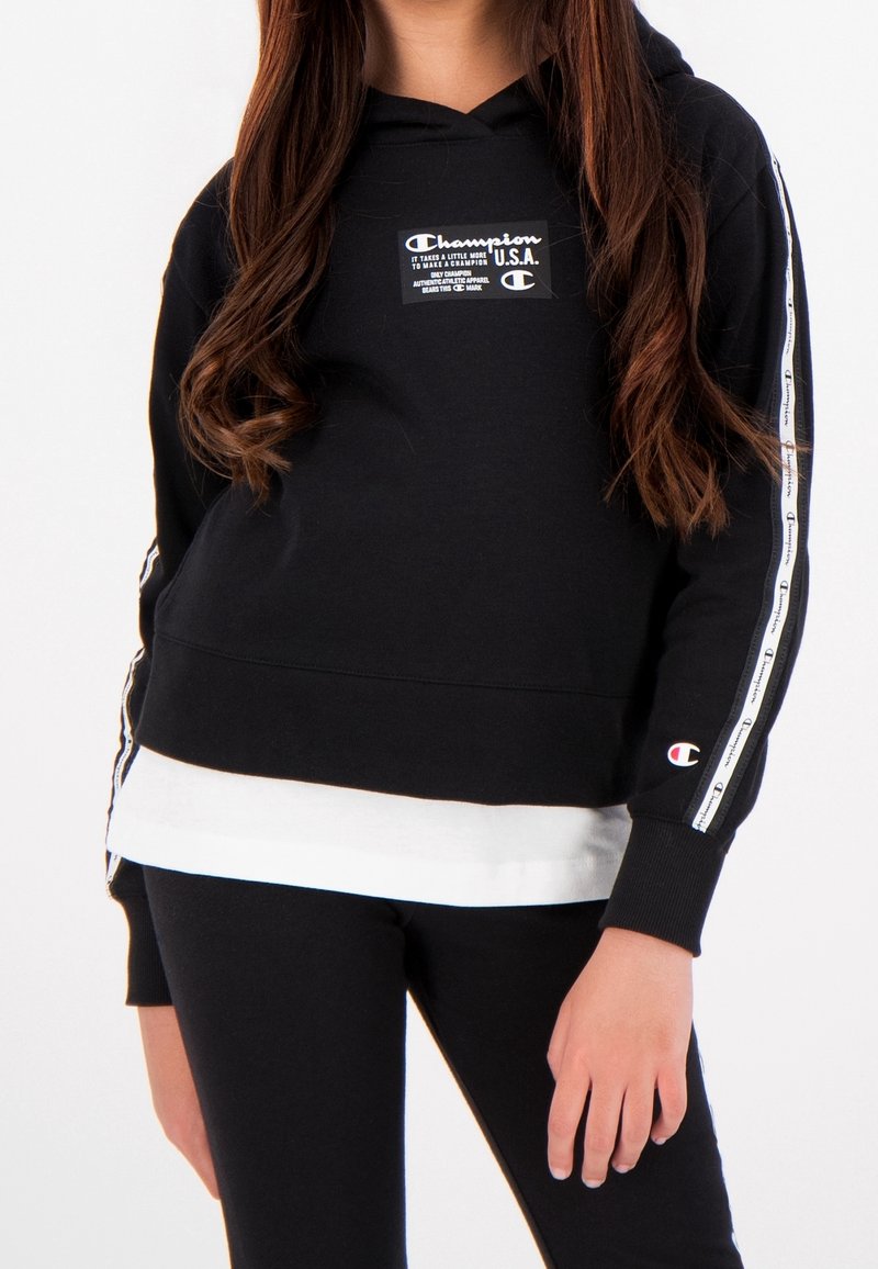 Haut à capuche noir Champion avec un patch logo sur le devant, des poignets côtelés et des bandes latérales comportant des noms de marque répétés. Associé à des leggings noirs.