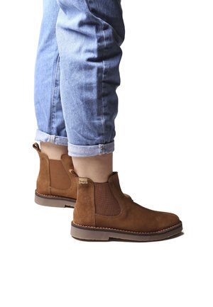 Bottines en suede marron avec des panneaux latéraux élastiques, des coutures détaillées et une semelle en caoutchouc plate, portées avec un jean bleu retroussé.