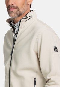 Beige zip-up jas met hoge kraag. Heeft een ritsluiting aan de voorkant, zijzakken en een logo patch op de mouw. Getextureerde stof.