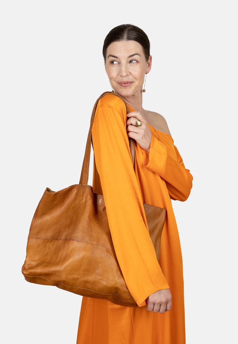 Bruine leren tote bag met een zachte textuur, ronde vorm en twee stevige hengsels. Weergegeven tegen een neutrale achtergrond.
