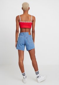 Person med kort blont hår som bär en röd crop top, blå denimshorts, vita Adidas-sneakers och randiga strumpor, som står mot en vit bakgrund.