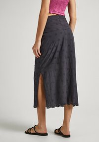 Falda gris hasta la rodilla con detalles de ojales florales y un dobladillo con forma de ondas. Presenta una abertura lateral y se combina con sandalias negras de tiras.