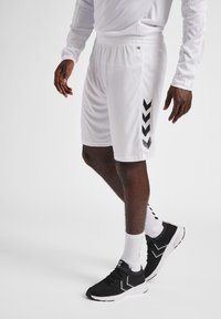 Shorts de sport blancs avec un motif en chevrons noirs sur le côté, fabriqués en tissu léger. Associés à des baskets noires et des chaussettes blanches.