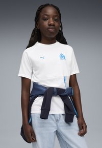 Balts sporta krekls ar zilo Marseļas logo un Puma zīmolu. Saskaņots ar gaiši zilām džinsām un tumši zilu sporta jaku, kas sasieta ap viduci.