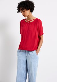 Top rosso a maniche corte con tessuto testurizzato, caratterizzato da un dettaglio del collo incrociato. Abbinato a jeans ampi blu chiaro.