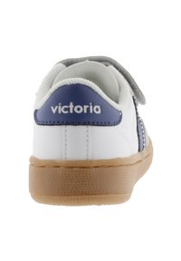 Sneaker bianco con tallone blu e cinturino grigio. Presenta suola in gomma testurizzata, parte superiore liscia con cuciture a contrasto e dettaglio con marchio.