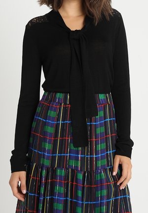 Haut en maille noire avec un détail à nouer au niveau du cou et des accents en dentelle. Associé avec une jupe à volants en tartan de couleur verte, bleue, rouge et noire.