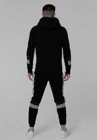 Felpa nera con collo alto e maniche aderenti, abbinata a pantaloni jogger neri con accenti colorati e branding lungo i lati.