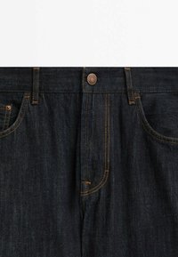 Tummat farkut, joissa on perinteinen viiden taskun muotoilu, kuparinvärinen nappi ja kontrastiväri keltainen ompelu saumoissa.