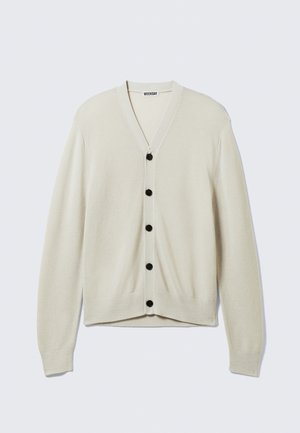 Beige cardigan med V-hals, langermet og svarte knapper foran. Strikket tekstur, tettsittende stil, glatt stoff. Merkeetikett synlig.