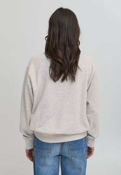 Šviesiai pilkas megztinis su laisvu kirpimu, nuleistais pečiais ir raukšlėtais rankogaliais, derinamas su mėlynomis džinsomis, turinčiomis subtilių siuvinėjimo detalių.