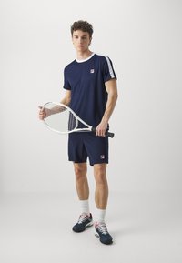 Tengerészkék sportöltözet, amely rövid ujjú, kerek nyakú pólóból, fehér részletekkel és hozzá illő sortból áll, teniszkészletet tartva.