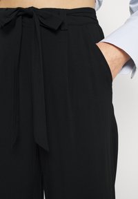 Svarta byxor med ett tygbälte knutet vid midjan, med sidofickor och en slät yta, beskurna för en åtsittande look.