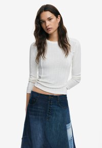 Weißes, geripptes Langarmshirt kombiniert mit einem Jeansrock, der mit einer Knopfleiste und einem Patchwork-Design in verschiedenen Blautönen versehen ist.