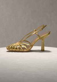 Χρυσό μεταλλικό σανδάλι με λουράκια και ψηλό τακούνι, με αγκράφα στον αστράγαλο και ανοιχτή μύτη, τοποθετημένο σε φωτεινή, στρογγυλή επιφάνεια πάνω σε ουδέτερο φόντο.